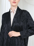 A close-up of a model wearing a black jacquard open-front jacket over a light beige collared shirt. The jacket features a subtle floral or damask pattern and is styled with a ring on the model's hand.