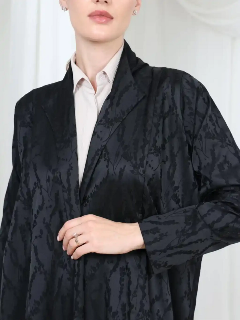 A close-up of a model wearing a black jacquard open-front jacket over a light beige collared shirt. The jacket features a subtle floral or damask pattern and is styled with a ring on the model's hand.