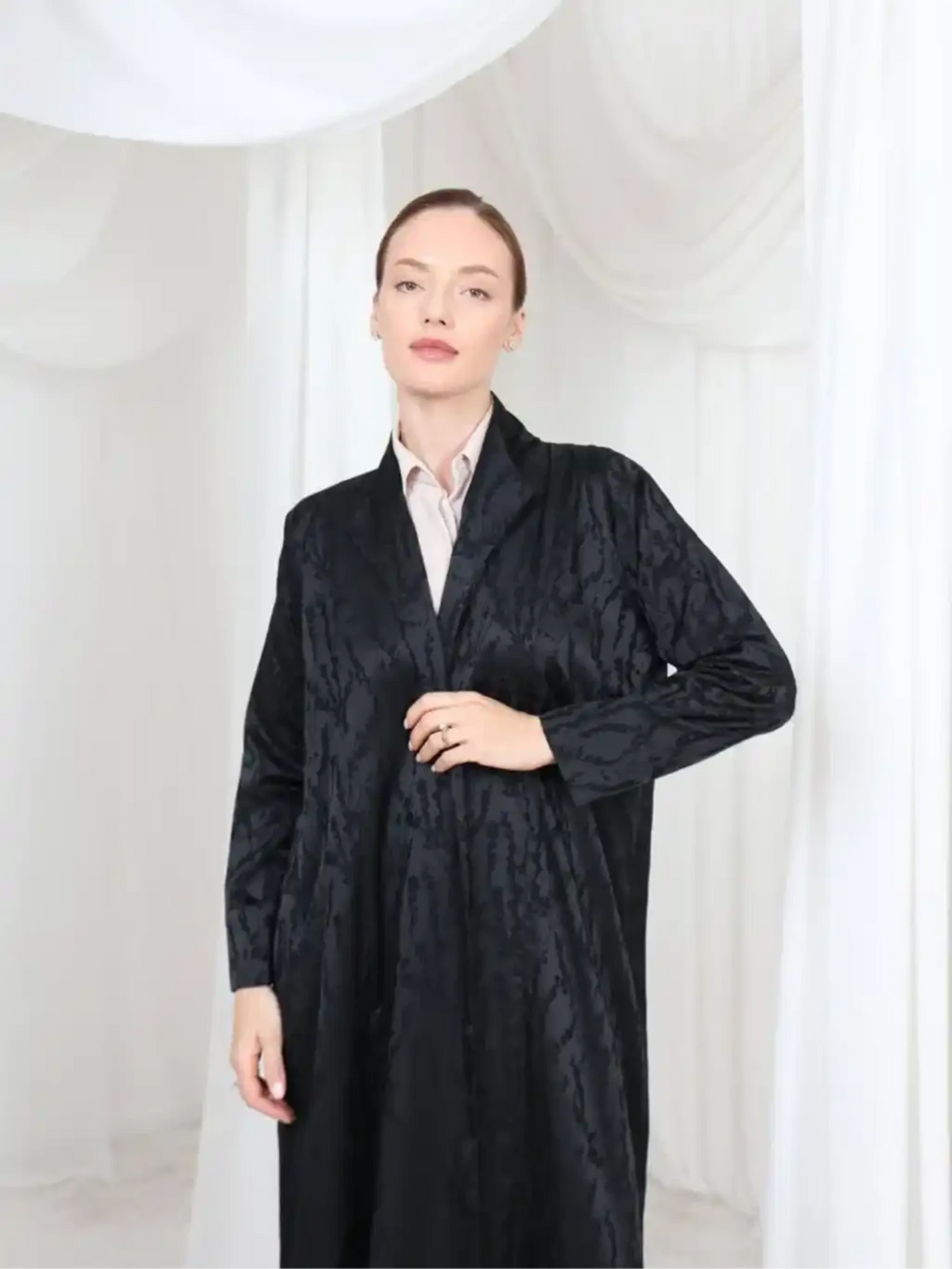 A woman modeling a long black textured coat over a light pink collared shirt, standing against a white draped fabric backdrop.