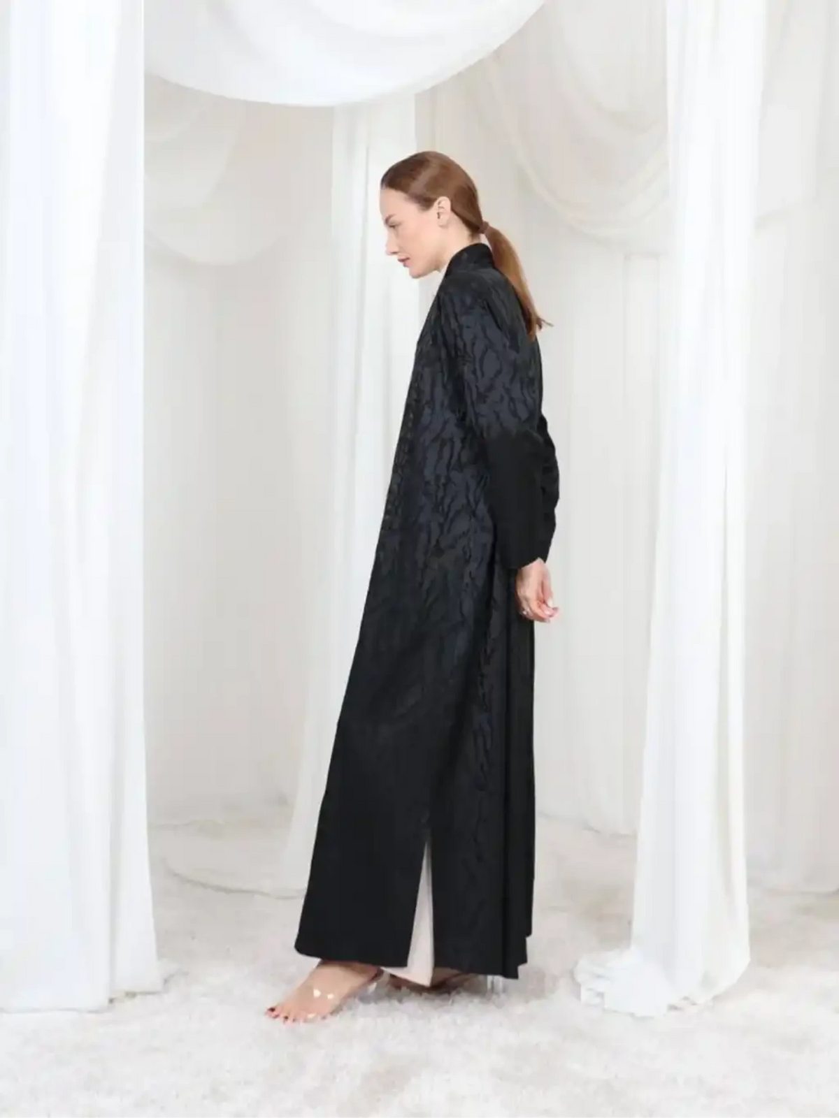 A woman wearing a long black textured abaya with a matching inner dress, standing in a white draped studio setting.
