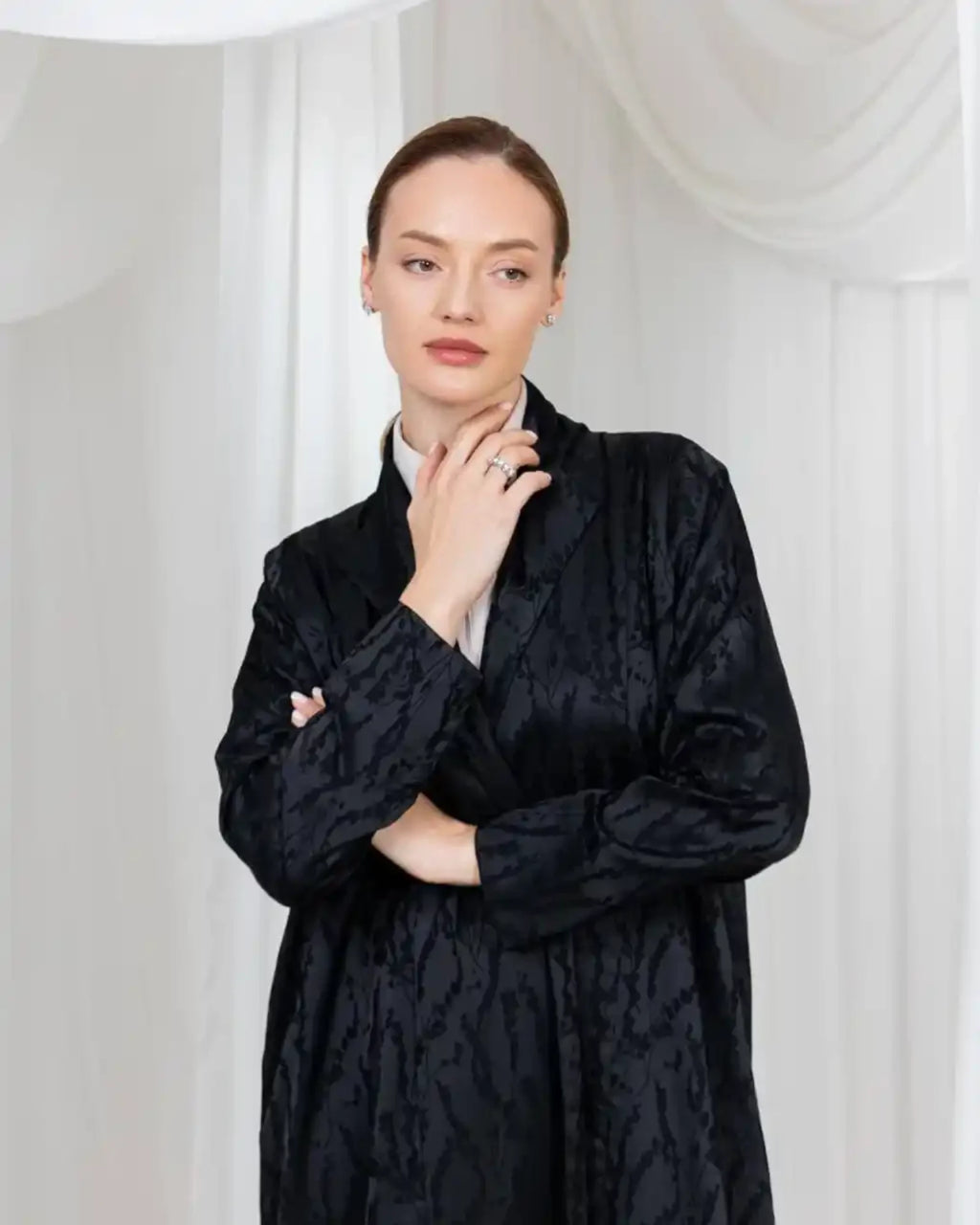 A woman modeling a black textured jacquard long-sleeve jacket over a white collared shirt, posed against a white draped fabric backdrop.