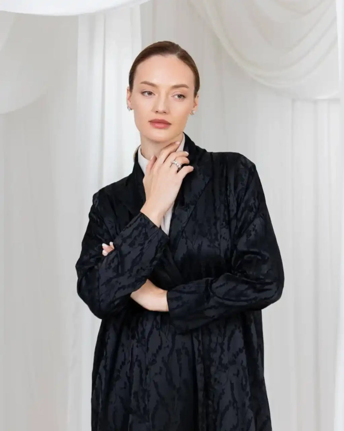 A woman modeling a black textured jacquard long-sleeve jacket over a white collared shirt, posed against a white draped fabric backdrop.