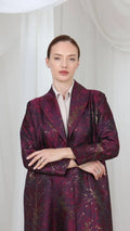 A woman stands against a white draped fabric background wearing a dark purple and burgundy patterned blazer over a light beige collared shirt.