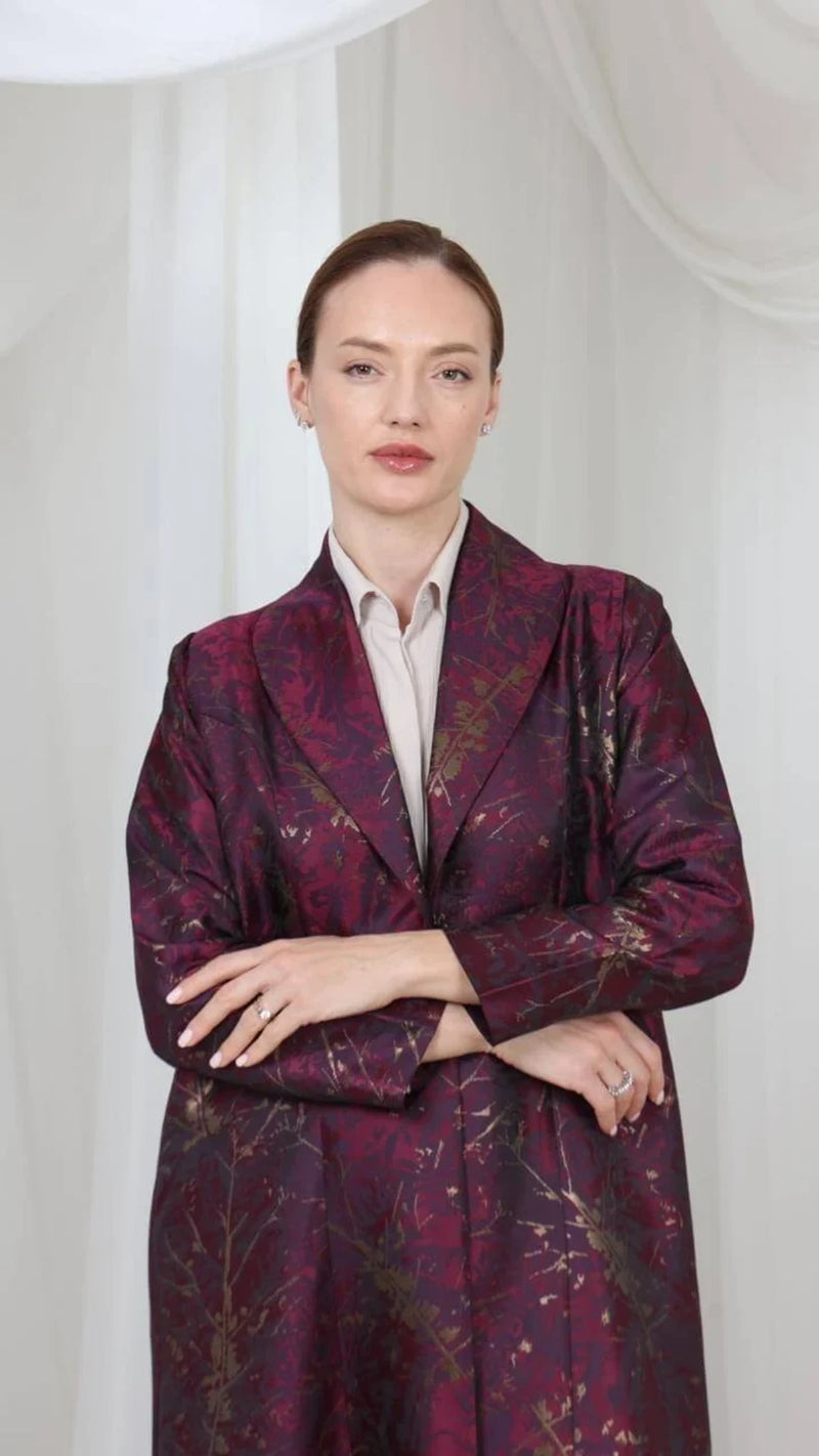A woman stands against a white draped fabric background wearing a dark purple and burgundy patterned blazer over a light beige collared shirt.