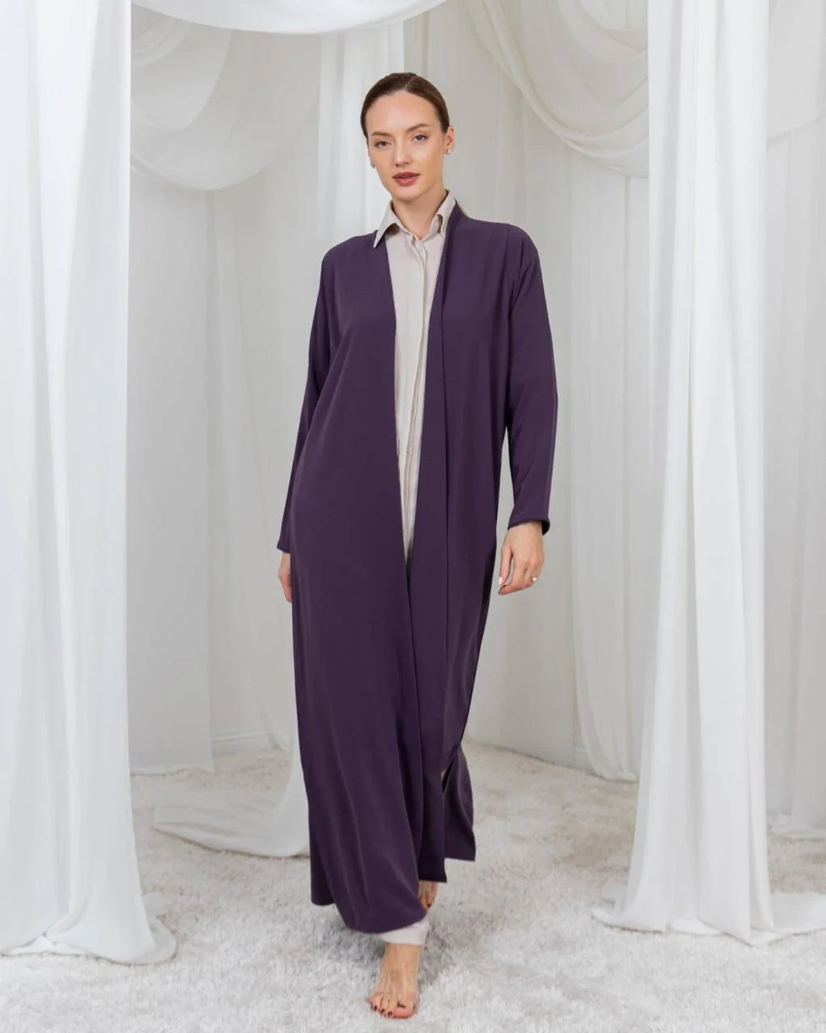 A woman modeling a long, flowing dark purple abaya over a light beige collared shirt, standing in a white draped studio setting.