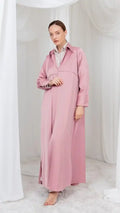 A woman modeling a long, dusty pink collared maxi dress with long sleeves and a relaxed fit, standing barefoot in a softly lit studio with white draped fabric and a white rug.