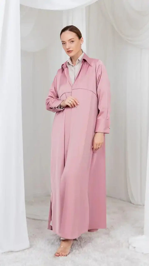 A woman modeling a long, dusty pink collared maxi dress with long sleeves and a relaxed fit, standing barefoot in a softly lit studio with white draped fabric and a white rug.