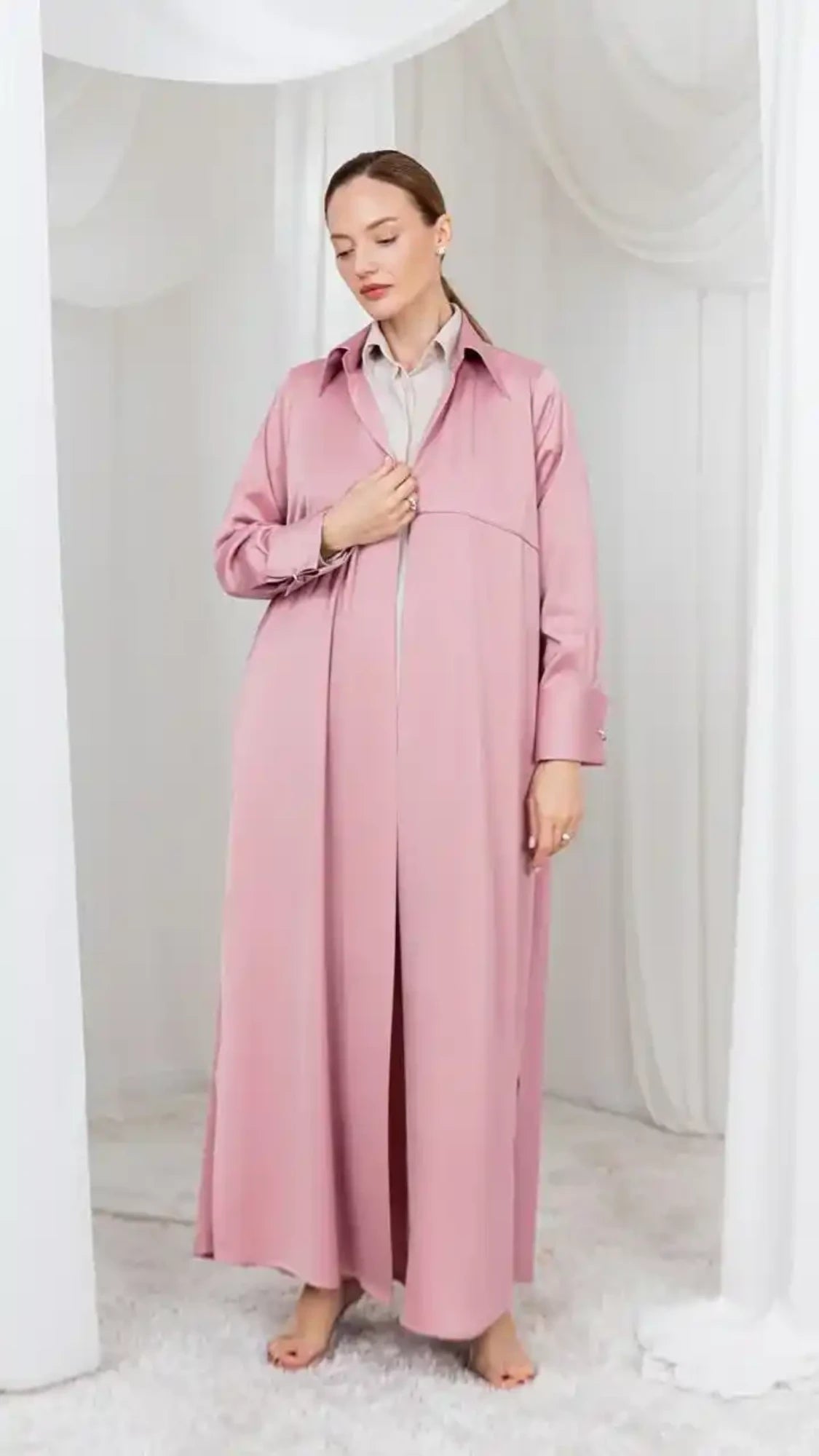 A woman wearing a long, dusty pink maternity trench coat over a white collared shirt, standing barefoot in a white draped studio setting.