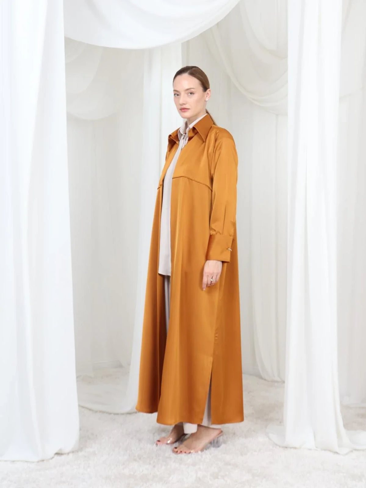 A woman stands barefoot in a studio draped with white fabric, wearing a long mustard-brown trench coat over a white top and wide-leg pants.