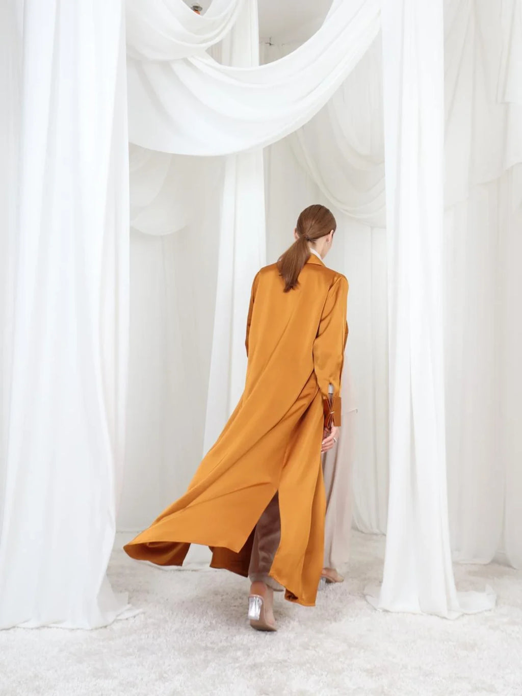 A woman viewed from behind wearing a flowing mustard-orange long coat over brown pants, standing in a white draped fabric setting.