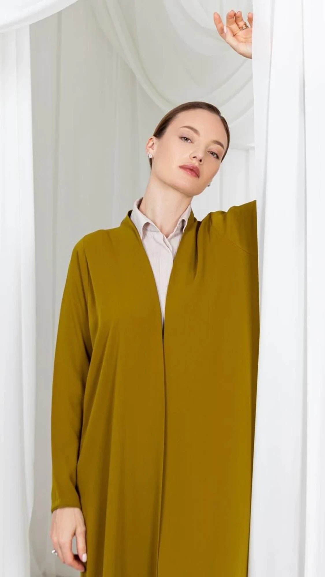 A woman wearing a long, flowing mustard yellow abaya over a light beige collared shirt, posing in front of white draped curtains.