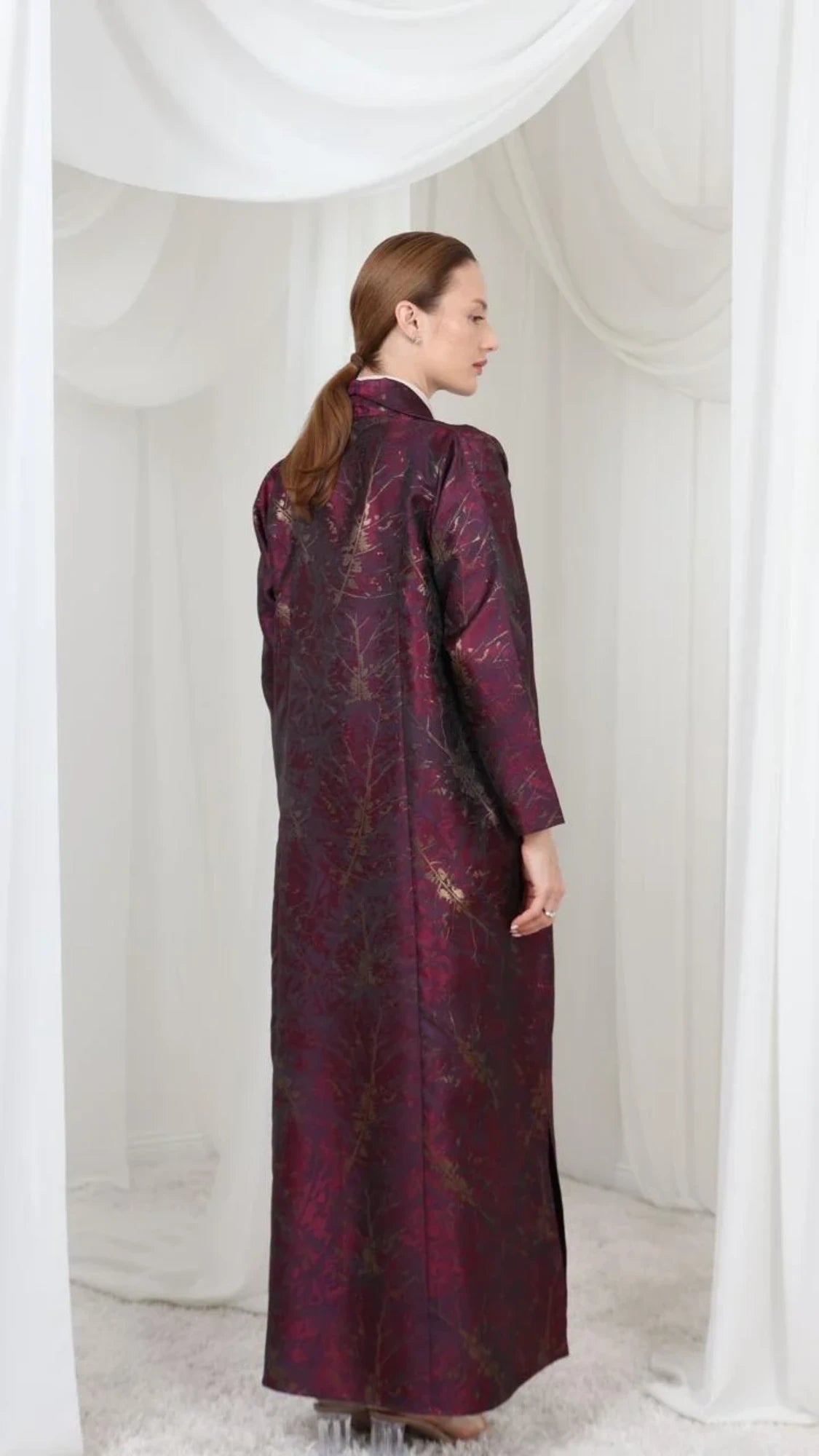 A woman wearing a long, dark plum purple robe with gold floral embroidery, photographed from behind in a white draped studio setting.
