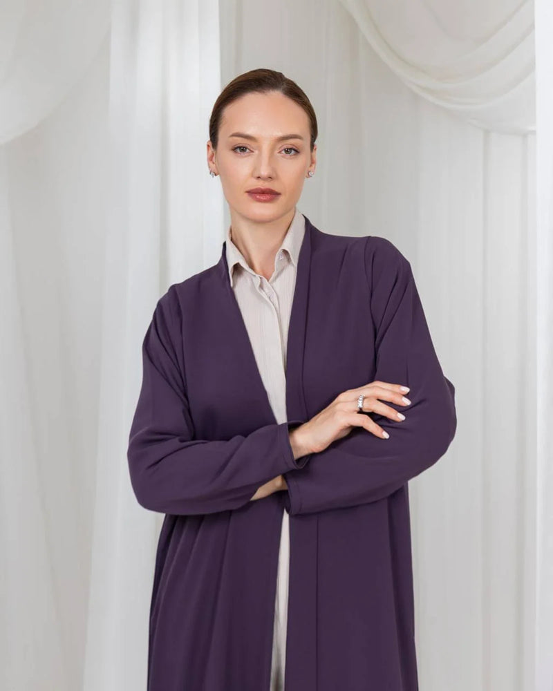 A woman modeling a long-sleeved, open-front purple cardigan over a light beige collared shirt, standing against a white draped curtain backdrop.